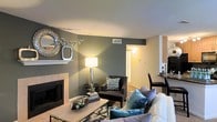 Located at 2904 Calvary Dr in Raleigh, NC, this single-floor residence offers a thoughtfully designed interior with a cohesive and contemporary aesthetic. The living area serves as the heart of the home, anchored by a tiled fireplace with a clean white mantle that provides a classic focal point. The space is adorned with deep slate-gray accent walls that contrast beautifully against neutral tones, creating a sophisticated and welcoming atmosphere. Warm ambient lighting from wall sconces and stylish table lamps enhances the cozy feel throughout. The living room is generously sized, offering comfortable seating arrangements and an open flow to adjacent areas of the home. A decorative mirror and carefully curated accents on the mantle add a personalized, polished touch. Visible through an open passageway, a dining area with bar-height seating extends the functional living space. The layout promotes a seamless connection between rooms, making the home feel open and well-proportioned. Neutral carpeting and smooth ceilings with integrated HVAC vents contribute to a clean, move-in-ready presentation throughout this charming Raleigh residence.