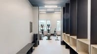 Located at 2-20 Malt Dr in Long Island City, NY, this expansive fitness center offers a comprehensive, well-appointed exercise facility across a single, generously sized floor. The space is designed with a modern aesthetic, featuring a dark industrial-style ceiling with abundant linear LED lighting that illuminates the entire facility, complemented by crisp white walls and bold black architectural framing details throughout.

The cardio zone is outfitted with a full row of treadmills, ellipticals, and stair climbers, all positioned alongside large windows that frame views of the surrounding Long Island City skyline, allowing natural light to fill the space during workouts. Adjacent to this area, a dedicated cycling room houses multiple Peloton-style spin bikes, complete with wall-to-wall mirrors and additional city views.

A functional training zone features a marked artificial turf lane equipped with a sled, battle ropes, plyometric boxes, and a cable/pull system, alongside a well-stocked dumbbell rack — ideal for high-intensity and athletic training. A strength and machine area provides a variety of cable machines and strength equipment reflected by floor-to-ceiling mirrors.

Rounding out the facility is a dedicated studio room with light hardwood flooring, mirrored walls, a ballet barre, and an open layout suitable for yoga, stretching, or group fitness classes. The overall design balances functionality with a clean, sophisticated ambiance.
