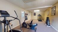 This single-room fitness center offers a functional space for cardiovascular and light strength training. The room features neutral beige walls, carpeted flooring, and a textured ceiling with flush-mounted fluorescent lighting supplemented by natural light from windowed entry doors on either end of the space. The layout accommodates multiple cardio machines, including a NordicTrack elliptical, a Schwinn upright stationary bike, a recumbent bike, and a treadmill positioned along the walls to maximize open floor space. A dumbbell rack with free weights provides options for strength training. Safety is well addressed, with a mounted fire extinguisher, an illuminated exit sign, and clearly posted Fitness Center Rules near the treadmill. A Lower Body workout guide chart is displayed on the wall, offering exercise references for users. Two entry doors—one windowed with a view of outdoor greenery—allow for ventilation and easy access. The overall atmosphere is straightforward and functional, providing residents with the essential equipment needed for a well-rounded workout routine in a clean, organized environment.