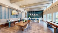 Located at 13611 Perry Rd in Houston, TX 77070, this single-story community clubhouse delivers a well-appointed gathering space designed for socializing and recreation. The open-plan layout flows seamlessly across wood-look plank flooring, connecting multiple distinct zones within one expansive interior.

The social hub centers on a sleek white kitchen island topped with blue upholstered barstools and backed by a striking teal tile feature wall, illuminated by pendant lighting. A sculptural multi-arm chandelier anchors the adjacent dining area, where a substantial wood communal table provides ample seating. A built-in banquette, clad in textured teal wallcovering and topped with a wall-mounted TV, offers a comfortable lounge nook.

The game area features a full shuffleboard table positioned alongside a generous bank of large windows and clerestory glazing above, flooding the space with natural light and offering views of the outdoor greenery. Exposed ceiling beams add architectural interest overhead.

A separate lounge zone presents soaring ceilings, white brick accent walls, and an arrangement of plush seating anchored by a patterned area rug, creating an inviting retreat. The cohesive coastal-contemporary palette of blues, whites, and warm wood tones unifies the entire space.