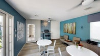 Located at 3101 Sweetwater Springs in Spring Valley, this single-floor business center offers a thoughtfully designed space that balances functionality with contemporary style. The interior is defined by bold teal accent walls that create a vibrant focal point, complemented by neutral tones throughout the rest of the space to maintain a professional yet welcoming atmosphere.

The room features a built-in workstation niche with a textured, decorative panel backdrop, providing a dedicated area for computer use. A striking starburst light fixture above the workstation adds an artistic element to the space. The lounge area is furnished with a modern taupe sofa and coffee table arrangement, ideal for informal meetings or relaxed work sessions.

High-top bistro tables with white chairs are positioned throughout the room, offering flexible seating options for individual work or small group gatherings. Wood-look plank flooring runs throughout the space, adding warmth and durability. A ceiling fan ensures comfortable airflow, while a glass-paneled door allows natural light to filter in from an adjacent area, enhancing the overall brightness of the space.