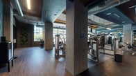 This fitness center offers a comprehensive workout environment designed with both functionality and aesthetics in mind. The space is organized into clearly defined zones, each serving a specific training purpose. The industrial-modern design is characterized by exposed ductwork running along the ceilings, concrete structural columns, and a dark ceiling palette, contrasted by warm wood-tone flooring and teal accent walls throughout.

The cardio zone is positioned along a wall of floor-to-ceiling glass windows, flooding the area with natural light and providing views of palm trees and the surrounding urban landscape. This section is outfitted with treadmills, ellipticals, stair climbers, and rowing machines. Wall-mounted flat-screen televisions are present for entertainment during workouts.

The strength training area features an extensive selection of selectorized cable machines and weight machines, complemented by a full rack of dumbbells and additional free-weight equipment. Large mirrors line the walls, enhancing the sense of space. A dedicated entry area with a teal accent wall, wall-mounted display shelving, and glass-paned doors separates zones while maintaining visual connectivity throughout the facility.