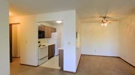 This single-floor residence offers a practical and comfortable layout that makes efficient use of its space. The living area is bright and welcoming, featuring neutral-toned carpeting and a ceiling fan that adds both style and airflow to the room. A half-wall partition subtly defines the transition between the living space and the kitchen, maintaining an open and connected feel throughout.

The kitchen is thoughtfully designed with a mix of upper and lower cabinetry in warm wood tones, providing ample storage for everyday needs. White appliances, including a refrigerator, electric range, and built-in microwave, are neatly integrated into the layout. The kitchen flooring features easy-to-clean tile or vinyl, contrasting with the carpeted living area. A pass-through counter along the partition wall enhances the flow between the two spaces, making it ideal for casual dining or entertaining.

The overall interior is finished in clean white walls that reflect light and create a sense of openness. The cohesive design elements throughout — from the cabinetry to the flooring transitions — give the space a uniform and well-maintained appearance. This residence is well-suited for those seeking a straightforward, move-in-ready home with a functional open-plan design.