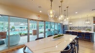 Located at 6801 Chesterbrook Ct in Raleigh, NC, this single-floor community amenity center offers a well-rounded selection of shared spaces designed for comfort, productivity, and wellness. The welcoming clubroom features warm hardwood-style flooring, a sectional leather sofa, accent chairs, and a patterned area rug, all bathed in natural light streaming through large floor-to-ceiling windows and sliding glass doors that open toward the outdoor pool area. Private office suites with glass-paneled walls and rich wood doors provide quiet workspaces just off the main lounge. The gourmet kitchen is a standout feature, showcasing a large quartz-topped island with seating, white cabinetry, a mosaic tile backsplash, stainless steel appliances, and statement pendant lighting. A connected dining area with an elegant marble-look table and additional pendant fixtures overlooks the pool through expansive glass doors. The fitness center is generously equipped with treadmills, stationary bikes, rowing machines, free weights, resistance equipment, and a mirrored strength training station, complemented by large windows with outdoor views and a water station. A dedicated package room with organized wall-mounted shelving rounds out the facility's practical amenities.