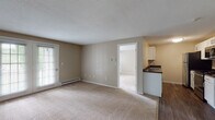 Located at 82 Brick Kiln Rd in Chelmsford, Massachusetts, this single-floor residence offers a well-organized layout designed for comfortable everyday living. The living area is finished with plush wall-to-wall carpeting and neutral beige walls, creating a warm and inviting atmosphere. A baseboard heating system runs along the perimeter, and a door with slatted blinds provides access to natural light and outdoor views. The space flows conveniently into the kitchen via a half-wall counter with a dark laminate surface, which functions as a natural divider. The kitchen is outfitted with crisp white cabinetry, dark countertops, and a full suite of stainless steel appliances including a refrigerator, range with electric cooktop, and an over-the-range microwave. Rich wood-tone flooring grounds the kitchen and extends into the bathroom. The bedroom is generously proportioned, featuring the same soft carpeting as the living area, a window with outdoor greenery views, and a baseboard heater. The full bathroom is clean and functional, featuring a tub-shower combination, a white vanity with ample cabinet storage, and wood-tone flooring, completing this practical and well-appointed residence.