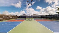 Located at 2507 Koji St in Mount Dora, Florida, this outdoor recreational facility features four dedicated pickleball courts arranged in a symmetrical layout. The courts are finished in a striking combination of blue and terracotta-red surfaces with clearly marked white boundary lines, providing excellent visibility during play. Each court is equipped with a regulation-height net, ensuring a proper playing experience for all skill levels.

The entire facility is enclosed by a sturdy black chain-link perimeter fence, which also incorporates windscreen panels to help reduce wind interference during play. Tall court lighting poles are positioned throughout the complex, allowing for evening matches and extended play after sunset. Seating benches are placed along the fence line, offering a comfortable resting area for players and spectators alike.

The courts are set within a residential neighborhood, with palm trees and landscaped grounds visible beyond the fencing, contributing to the characteristically sunny, Florida ambiance. A gravel border runs along the interior base of the fence, framing the playing surfaces neatly. Under the expansive Florida sky, the facility offers an open-air environment ideal for both casual recreational play and more competitive matches, making it a well-appointed community sports amenity in Mount Dora.