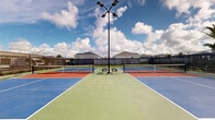 Located at 2507 Koji St in Mount Dora, Florida, this outdoor recreational facility features four dedicated pickleball courts arranged in a symmetrical layout. The courts are finished in a striking combination of blue and terracotta-red surfaces with clearly marked white boundary lines, providing excellent visibility during play. Each court is equipped with a regulation-height net, ensuring a proper playing experience for all skill levels.

The entire facility is enclosed by a sturdy black chain-link perimeter fence, which also incorporates windscreen panels to help reduce wind interference during play. Tall court lighting poles are positioned throughout the complex, allowing for evening matches and extended play after sunset. Seating benches are placed along the fence line, offering a comfortable resting area for players and spectators alike.

The courts are set within a residential neighborhood, with palm trees and landscaped grounds visible beyond the fencing, contributing to the characteristically sunny, Florida ambiance. A gravel border runs along the interior base of the fence, framing the playing surfaces neatly. Under the expansive Florida sky, the facility offers an open-air environment ideal for both casual recreational play and more competitive matches, making it a well-appointed community sports amenity in Mount Dora.