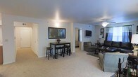 This single-floor residence showcases an open-concept design that seamlessly connects the dining and living areas, creating a spacious and functional layout. The interior is finished with soft, neutral-toned carpeting that extends throughout the main living spaces, complemented by clean white walls that enhance the sense of openness and light.

The dining area features a dark-toned bar-height table with matching stools, positioned beneath a framed wall mirror that adds depth and visual interest to the space. A hallway is accessible nearby, leading to additional rooms within the residence.

The living area is well-appointed with comfortable seating, including an armchair and sofa arranged around a coffee table atop a textured area rug. A wall-mounted flat-screen television serves as a focal point in this zone, while a ceiling fan with an integrated light fixture provides both air circulation and ambient lighting overhead.

The open sightlines between the two zones make the overall space feel expansive and cohesive. The consistently neutral color palette across the flooring, walls, and ceiling contributes to a calm, inviting ambiance throughout the home.