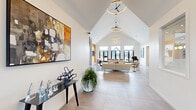 This sophisticated single-floor amenity space offers a compelling blend of modern architecture and thoughtful design. The welcoming entrance features a vaulted, pitched-glass entryway with a sleek pendant light, setting an elegant tone from the moment of arrival. Large-format tile flooring extends throughout, unifying the space seamlessly.

The expansive main lounge commands attention with its dramatic cathedral ceiling adorned with circular pendant lights and ceiling fans. A striking floor-to-ceiling dark tile fireplace serves as a bold focal point, flanked by large-scale artwork and natural wood wall accents. Generous floor-to-ceiling windows line the far wall, flooding the space with natural light and offering outdoor views. The lounge accommodates comfortable seating arrangements alongside a foosball table, making it ideal for both relaxation and social gatherings.

The kitchenette area is designed for functionality and style, featuring sage green cabinetry, a white quartz-topped island with bar seating, globe pendant lighting, dark tile backsplash, open wood shelving, and stainless steel appliances. A beverage and coffee station adds everyday convenience, and a glazed door opens directly to an outdoor area.

A corridor leads to additional private rooms, completing this well-appointed and visually cohesive community space.