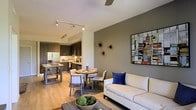 This thoughtfully designed single-floor residence showcases a seamless open-concept layout that unifies the living, dining, and kitchen spaces into one cohesive and inviting environment. The living area features a clean, modern aesthetic with a neutral-toned sofa accented by bold blue throw pillows, anchored by a rustic wood coffee table and complemented by a decorative bowl centerpiece. A striking accent wall in warm gray provides a sophisticated backdrop for contemporary wall art and sculptural décor pieces, adding visual depth and character to the space. The dining area flows naturally between the living room and kitchen, centered around a round table with cushioned chairs that invite comfortable gatherings. The kitchen is a standout feature, boasting dark cabinetry with modern hardware, stainless steel appliances, and a light countertop that brightens the space. A kitchen island with bar seating offers additional functionality and casual dining options. Track lighting runs across the ceiling, providing flexible, targeted illumination throughout, while light wood-toned flooring ties the entire open space together with warmth and continuity. The overall ambiance is contemporary, polished, and welcoming.