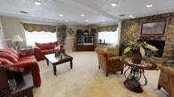 This single-floor space centers around a generously sized living area that combines traditional design elements with warm, inviting details. The room is finished with neutral wall-to-wall carpeting that adds softness underfoot, while a textured popcorn ceiling with recessed lighting provides even, ambient illumination throughout.

A standout architectural feature is the floor-to-ceiling natural stone accent wall on one side of the room, adding rustic character and visual depth. Multiple windows dressed with decorative scalloped valances in muted green tones allow natural light to fill the space, creating a bright and airy atmosphere.

The living area is furnished with a combination of seating options, including a bold red sofa, patterned accent chairs, and light-toned armchairs, arranged around a dark wood coffee table. A traditional wooden entertainment armoire serves as a central media focal point. Decorative greenery and potted plants are placed throughout, adding natural elements and enhancing the room's warmth. The overall ambiance is comfortable and classic, suggesting a communal or shared living environment suited for relaxed gathering and everyday use.