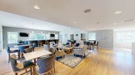 Located at 969 W Main Rd in Middletown, Rhode Island, this two-floor space offers a well-appointed layout designed for both productivity and wellness. The upper floor serves as the main gathering area, highlighted by a combined living and dining room with soaring ceilings, gleaming light hardwood floors, and an abundance of natural light streaming through multiple white-trimmed windows. This expansive open-plan area features comfortable lounge seating, dining tables with upholstered chairs, and a granite-topped bar counter with exterior deck access — ideal for collaborative or social use. Curated artwork and a neutral warm-gray palette lend a polished yet inviting atmosphere throughout. A café nook with cabinetry and a mounted TV adds further functionality. The two private offices provide dedicated workspace, one of which features a distinctive reclaimed wood accent wall and built-in storage. A connecting hallway flows seamlessly between areas. The lower floor houses a fully equipped exercise room with rubber flooring, cardio machines, a cable machine, free weights, wall-length mirrors, and a wall-mounted mini-split system — providing a comprehensive fitness facility. Recessed lighting and multiple windows ensure the workout space remains bright and airy.