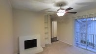 This single-story home in Bedford, Texas offers a comfortable and functional living space. The main living area serves as the focal point of the home, featuring a white brick fireplace that adds a classic, cozy element to the room. Adjacent to the fireplace is a set of built-in shelving units, providing convenient display and storage space. A ceiling fan with a light fixture is centered overhead, promoting air circulation and adding to the room's practicality. The flooring in the living area is carpeted, contributing to a warm and inviting atmosphere. Neutral-toned walls complement the light color palette throughout, creating a clean and versatile backdrop. A textured ceiling adds subtle architectural character. Natural light filters in through what appears to be a sliding glass door or large window along one wall, brightening the space. A hallway visible in the background leads to additional areas of the home, with contrasting darker flooring suggesting a transition into other rooms. Overall, the space presents a straightforward, well-maintained interior well-suited for comfortable everyday living.