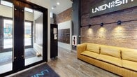 Located at 1030 Fifth Ave in Pittsburgh's dynamic urban core, this single-floor lobby space showcases a striking industrial-modern aesthetic that masterfully balances historic architectural elements with contemporary design. The entrance greets visitors with wood-look flooring, a bold branded signage wall, and a welcoming yellow leather sofa positioned against exposed brick, setting an inviting yet sophisticated tone.

The lobby opens into a generously proportioned common area distinguished by soaring ceilings that amplify the sense of space. Exposed brick walls and visible ductwork pay homage to the building's industrial roots, while a dramatic cluster of copper pendant lights serves as a striking focal point overhead. High-top tables with industrial-style stools provide casual seating, complemented by a built-in coffee station with cabinetry nearby.

A feature wall finished with geometric dark-gray paneling anchors a mounted flat-screen television, adding a modern, polished contrast to the raw materials surrounding it. An interior mezzanine level with black metal railings overlooks the main floor, reinforcing the vertical drama of the space. Corridors lined with clean white walls and accent brick lead to additional building access points, maintaining design continuity throughout.