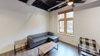 This single-floor unit showcases a compelling blend of modern and industrial design elements. The living area serves as the social hub of the space, featuring dramatically dark exposed ceilings with visible ductwork and structural beams, contrasted against crisp white walls. Wood-look flooring runs throughout, and large wood-framed windows allow natural light to fill the room while offering views of the surrounding building exterior. Ceiling fans are present in multiple rooms for added comfort.

The kitchen is well-appointed with white shaker-style cabinetry, white subway tile backsplash, quartz countertops, and slate-finish appliances including a full-size refrigerator, electric range, and over-the-range microwave. Industrial-style pendant lighting complements the dark exposed ceiling above.

Each bedroom is functional and compact, equipped with a ceiling fan, wood-look flooring, a wall-mounted television, and a built-in desk workspace. Select bedrooms feature windows with wood-framed trim, while at least one includes an open closet with built-in shelving and hanging rods.

The bathrooms maintain a clean, minimalist aesthetic with all-white walls, wood-look tile flooring, white vanities, and sleek black hardware accents throughout.