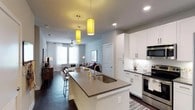 Located in the desirable Steiner Ranch community of Austin, Texas, this single-story residence offers a thoughtfully designed open-concept floor plan that seamlessly connects the kitchen, dining, and living areas. The kitchen is a standout feature, showcasing white shaker cabinetry, stainless steel appliances, a subway tile backsplash, and a large center island with a quartz countertop and undermount sink — perfect for both cooking and casual dining. Bold cylindrical pendant lights in a warm gold hue add a modern, stylish focal point above the island. Dark hardwood floors run throughout the main living space, providing a rich contrast to the light gray walls and white trim. Recessed lighting and large windows dressed with sheer curtains allow natural light to fill the space, creating a bright and welcoming ambiance. The living area offers generous room for comfortable furnishings and flows naturally from the kitchen, making the layout ideal for everyday living and entertaining. The overall design balances contemporary aesthetics with functional living, featuring clean lines, modern fixtures, and a neutral color palette that lends itself to a variety of personal styles.