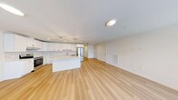 Located at 7 Arbutus Path in Mount Sinai, NY, this thoughtfully designed 2-bedroom, 2-bathroom single-story home offers a clean, modern aesthetic with an efficient and comfortable layout. The heart of the home is a generous open-concept living, dining, and kitchen area, unified by warm light hardwood flooring that extends throughout. The kitchen is a standout feature, showcasing crisp white shaker cabinetry with matte black hardware, marble-style tile backsplash, quartz countertops, stainless steel appliances including a French door refrigerator and electric range, and a central island that provides additional prep and seating space. Recessed and pendant lighting complete the polished look. The entry hallway welcomes natural light through multiple windows, creating a bright and airy arrival experience. Both bedrooms are carpeted for comfort, with the spacious primary bedroom featuring large windows and a sliding glass door that opens to views of lush greenery outdoors. The bathrooms are finished with marble-look tile flooring, white vanities, and backlit mirrors for an elegant touch. Additional conveniences include four closets, a dedicated laundry room, and a utility room, ensuring ample storage throughout this well-appointed home.