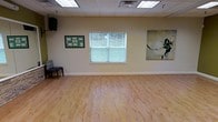 This single-floor studio space offers a clean, open layout designed for versatility and functionality. The room is finished with smooth, light-toned hardwood flooring that adds warmth and is well-suited for movement-based activities such as dance, fitness, or yoga classes. A drop-tile ceiling with recessed fluorescent lighting ensures consistent, even illumination throughout the space. A centrally positioned window fitted with horizontal blinds allows natural light to filter in while maintaining privacy, offering a pleasant view of the surrounding greenery. The walls are painted in a neutral warm beige, creating a calm and welcoming atmosphere. Decorative touches include a large botanical-inspired canvas artwork on one wall and a framed bulletin board, adding character and a sense of community to the environment. A wall-mounted speaker is present, supporting audio needs for classes or events. A single chair is positioned along one wall, hinting at a reception or waiting area. The overall ambiance is bright, airy, and functional — well-suited for professional or wellness-oriented uses.