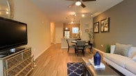 This single-story space showcases an open-concept floor plan that connects the living, dining, and kitchen areas into one cohesive and functional layout. Hardwood-style flooring flows throughout, lending warmth and visual continuity across the space. The living area features a ceiling fan with integrated lighting, while a track lighting system extends toward the dining and kitchen zones, providing layered illumination throughout.

The dining area comfortably accommodates a round table with upholstered chairs, positioned beneath pendant lighting that adds a focused, decorative touch. Adjacent to the dining space, the kitchen features a peninsula-style counter with bar seating, stainless steel appliances, and a window that introduces natural light. The neutral wall tones are accented by a deeper olive-green feature wall in the living area, creating visual contrast and depth. Framed wall art further enhances the area's character.

A hallway visible from the main living space leads to additional rooms, suggesting a well-organized layout. The overall ambiance is warm, inviting, and thoughtfully designed for both everyday living and entertaining.