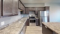 Located at 3031 Roosevelt Rd in Saint Cloud, Minnesota, this single-floor residence offers a well-designed, functional layout with modern finishes throughout. The kitchen serves as a central highlight, showcasing rich dark-toned cabinetry, granite-style countertops, and a full suite of stainless steel appliances including a refrigerator, electric range, and dishwasher. A center island provides additional prep space and enhances the kitchen's utility, while light wood-look flooring grounds the area with a clean, contemporary feel. The kitchen opens directly into a generously proportioned living area finished with plush carpeting and recessed lighting, creating a comfortable and inviting atmosphere. Large windows and a sliding glass door in the living space allow natural light to pour in and provide access to a private outdoor balcony with exterior views. The bathroom is finished with stone-patterned tile flooring, a large vanity with an integrated sink, an oversized mirror, and accessibility-friendly grab bars. A dedicated in-unit laundry area accommodates a full-size washer and dryer, adding everyday convenience. Baseboard heating is present throughout, ensuring comfort during Minnesota's colder seasons. Overall, the space combines practical amenities with modern design elements in a cohesive, move-in-ready package.
