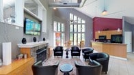 This single-floor community space offers an inviting and well-appointed lounge area designed to bring people together in comfort. The room immediately impresses with its dramatic vaulted ceilings, which soar overhead and are complemented by large, floor-to-ceiling windows that flood the interior with an abundance of natural light and offer views of the surrounding greenery. Exposed ductwork and a ceiling fan add an industrial yet approachable character to the space.

At the heart of the lounge sits a striking stone fireplace, providing a warm focal point around which comfortable black leather club chairs are thoughtfully arranged. Small accent tables are positioned throughout, creating an ideal setting for conversation or relaxation. A neutral-toned area rug anchors the seating arrangement, adding warmth to the tile flooring.

Adjacent to the main lounge area, a kitchenette is equipped with cabinetry, a countertop bar, and appliances, making it practical for gatherings and events. A wall-mounted flat-screen television near the fireplace adds to the entertainment offerings of the space. A bold accent wall in deep red provides a vibrant contrast to the soft grey tones of the surrounding walls, lending the space a lively and modern energy throughout.