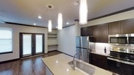 This single-floor space showcases a thoughtfully designed open-concept layout that seamlessly connects the kitchen and living areas. The kitchen serves as the centerpiece, featuring dark espresso-toned cabinetry that provides a striking contrast against light-colored quartz countertops. A large kitchen island with an undermount sink and a sleek arc faucet offers both functionality and a natural gathering point. Stainless steel appliances, including a full-size refrigerator, contribute to the modern aesthetic, while under-cabinet lighting illuminates the workspace effectively.

Three cylindrical pendant lights hang at varying heights above the island, adding a contemporary design element while providing focused task lighting. Recessed lighting throughout the space ensures consistent, bright illumination. Rich dark hardwood flooring runs throughout, grounding the space with warmth and continuity.

Adjacent to the kitchen, the living area benefits from natural light streaming through a set of elegant French doors, which also provide direct outdoor access. Built-in shelving along one wall offers practical storage and display opportunities. Dark crown molding frames the white ceilings, adding a refined architectural detail throughout the space.