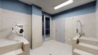 This single-floor space appears to be a purpose-built utility or pet grooming room, designed with practicality and cleanliness at its core. The walls feature large-format beige ceramic tiles on the lower half, transitioning to a muted steel-blue painted finish on the upper portion, creating a clean, two-tone aesthetic that is both visually organized and easy to maintain. The floor is covered in small mosaic tiles, providing a slip-resistant surface well-suited for wet conditions.

The room is equipped with built-in raised washing stations or grooming platforms positioned along the sides, constructed from matching tiled material to ensure durability and hygiene. A wall-mounted utility attachment, likely a dryer or sprayer arm, is visible on one side, indicating the space is outfitted for professional or dedicated grooming use.

A glass-paneled interior door leads to an adjacent area, while a solid gray door provides access to another room or exit, offering convenient flow through the space. Overhead fluorescent lighting ensures bright, even illumination throughout. The overall design prioritizes function, cleanliness, and efficiency, making this a well-considered utility space.