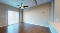 This single-floor unit showcases a distinctive industrial-contemporary design highlighted by rich, stained concrete floors that run throughout the entire space, adding warmth and visual depth. The open-concept living area features soaring ceilings with an exposed cylindrical HVAC duct serving as a bold architectural detail, complemented by a ceiling fan and vertical blind-covered sliding door that allows for natural light control. The kitchen is a standout feature, boasting tall white shaker cabinetry extending nearly to the ceiling, a light-toned quartz peninsula with an undermount double sink, a subway tile backsplash, and a full suite of stainless steel appliances including a refrigerator, range, and microwave. Recessed lighting illuminates the kitchen and dining area, enhancing the open, airy feel. The bedrooms are generously sized, each offering natural light through horizontal-blind windows, with one room featuring a distinctive structural column. The bathroom includes a glass-enclosed walk-in shower with white subway tile, a white vanity with ample storage, and consistent stained concrete flooring. Throughout, neutral warm-gray walls provide a cohesive, modern backdrop suitable for a variety of interior styles.