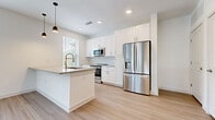 This thoughtfully designed 1-bedroom, 1-bathroom unit offers a cohesive and modern living experience across a single floor. The open-concept kitchen and living area serves as the heart of the space, featuring crisp white shaker-style cabinetry that extends to the ceiling for ample storage, a classic hexagonal tile backsplash, and a suite of stainless steel appliances including a French door refrigerator, electric range, and built-in microwave. A peninsula with a stainless steel sink and light countertops provides additional prep space and casual seating potential, while stylish pendant lights add a contemporary finishing touch. Light-toned hardwood-style flooring flows throughout the main living area, complementing the neutral white walls and creating a bright, airy atmosphere. Multiple large windows with horizontal blinds invite abundant natural light and outdoor views into the living room, which also features a glass-paned entry door. The bedroom is carpeted for comfort, offers a generous window with outdoor views, and includes access to a dedicated closet. The bathroom is finished with light wood-style flooring and features a sleek, glass-enclosed shower adorned with striking marble-patterned tile walls, conveying a sense of refined elegance.