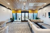 Located at 217 S Weber St in downtown Colorado Springs, this well-appointed lounge space offers a thoughtfully designed interior that blends comfort with contemporary style. The standout architectural feature is the warm wood-slat ceiling, which spans the entire space and adds a natural, textured contrast to the clean white walls and modern finishes throughout.

The main lounge area is anchored by a generously sized sectional sofa arrangement upholstered in neutral tones of cream, gray, and blue, centered on a patterned carpet that adds visual depth to the floor. A wall-mounted flat-screen television with an integrated soundbar serves as a focal point, flanked by curated artwork and complemented by a built-in beverage station with a wine refrigerator. Recessed lighting and potted greenery add to the welcoming ambiance.

A more intimate seating nook provides a quieter retreat, furnished with patterned accent chairs and an ottoman, with its own wall-mounted display screen — ideal for small gatherings or private use.

The space opens directly onto a spacious balcony through full-height glass doors, framed by bold yellow valances. The balcony features tiled flooring, glass railings, and bistro-style high-top tables, offering elevated views of the Colorado Springs skyline and the surrounding mountain backdrop — a striking complement to this refined interior space.