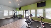 This fitness center is thoughtfully designed to accommodate a full range of workout activities across distinct functional zones. The space features vaulted ceilings that rise to an upper mezzanine level, decorated with plants and framed by a dark-railed balcony, lending the facility a sense of openness and vertical scale. The flooring transitions between durable rubber matting in the workout zones and wood-look hardwood along the central corridor, providing both practicality and aesthetic appeal.

The cardio zone houses a treadmill, elliptical, and stationary bikes positioned on dark rubber flooring, complemented by a wall-mounted television for entertainment during workouts. Adjacent to this area, the strength training zone features a multi-functional cable machine, a weight bench, a dumbbell rack, and additional free weights, all set against a bold accent wall in vibrant green that enlivens the space. A large mirror spans one wall to assist with form and technique.

Multiple windows fitted with white blinds allow generous natural light throughout, while a water dispenser station provides convenient hydration access. The central corridor leads to a glass-paneled entry door, offering exterior views and enhancing the bright, welcoming atmosphere of the facility.