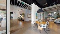 Located on Old Brick Rd in Glen Allen, Virginia, this thoughtfully designed single-floor commercial space blends contemporary aesthetics with highly functional work environments. The interior is defined by exposed black ceiling infrastructure contrasted against white structural elements and polished concrete flooring, creating an industrial-modern atmosphere that feels both creative and professional.

The entry area is welcoming and open, featuring floor-to-ceiling windows that flood the space with natural light and offer views of the surrounding streetscape. Sculptural pendant lighting and dark vertical slatted partitions elegantly define zones without sacrificing openness. A built-in bench seating area near the entrance provides a casual gathering spot.

Deeper into the space, multiple distinct zones cater to varied work styles. Open collaborative areas feature round and rectangular tables with mixed seating. A lounge area with tufted green sofas, curated artwork, and warm accent lighting offers a relaxed setting. Private focus nooks with built-in desks and pegboard backdrops provide quiet individual workspaces.

A well-appointed kitchenette includes white tile backsplash, open wood shelving, a large island with bar seating, and globe pendant lights. A secondary prep area features light-blue subway tile, open shelving, and under-counter refrigeration. Glass-partitioned conference rooms and semi-private meeting areas with large windows complete this versatile, design-forward workspace.