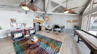 This single-floor space makes a strong first impression with its dramatic vaulted ceiling, where exposed white-painted wood beams and visible ductwork create an industrial-chic aesthetic. Ceiling fans and a statement woven pendant light hang from the apex, adding both function and character to the lofty interior.

The open-plan layout seamlessly integrates multiple zones. A living area anchors the space with a bold, multicolored area rug and a mid-century style oval coffee table surrounded by modern accent chairs. Adjacent to this, a dining area features a round marble-top table with sleek dark chairs, positioned near a striking arched brick fireplace that serves as a natural focal point along the wall.

A fully equipped kitchenette lines the back wall, outfitted with wooden cabinetry, stainless steel appliances, and a raised counter with bar seating. Light wood-toned flooring runs throughout, unifying the various zones cohesively.

A "PLAY" wall sign adds a playful, welcoming touch to the overall ambiance. The space balances energetic, colorful design elements with functional areas, making it well-suited for social gatherings or community use.