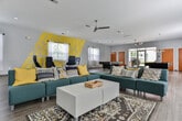 Located at 316 Columbia Drive in Carrollton, Georgia, this single-floor community clubhouse serves as a dynamic social hub for the Timbercrest community. The space is designed with an open, flowing layout that encourages gathering and recreation, finished throughout with light wood-look flooring that ties the various zones together seamlessly.

The main lounge area is generously proportioned, featuring a large sectional seating arrangement in teal upholstery accented with bold yellow and navy throw pillows, centered around a white coffee table. A wall-mounted flat-screen television anchors one end of the lounge, while sheer white curtains on multiple windows allow ample natural light to fill the room. A branded Timbercrest step-and-repeat backdrop adds a community-identity focal point to this area.

Adjacent to the lounge is a stylish conversation nook defined by a bold geometric yellow and gray accent wall mural. Navy modern lounge chairs and a round white side table create an intimate seating vignette, adding visual contrast and energy to the space. Ceiling fans throughout maintain comfort across the open floor plan.

The game room houses two full-size billiard tables illuminated by pendant lights, with direct access to the exterior through glass-paneled doors that bring in views of the surrounding greenery. Additional seating and storage areas round out the space, making this clubhouse a well-appointed and inviting community amenity.