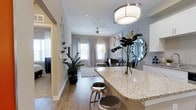 This 1-bedroom, 1-bathroom residence is designed with a modern, open-concept layout that maximizes the sense of space across its single floor. The kitchen serves as a central hub, featuring sleek white cabinetry, granite countertops with an extended breakfast bar, and stylish pendant lighting overhead — ideal for both casual dining and entertaining. Durable hardwood-style flooring flows seamlessly through the main living areas, adding warmth and continuity throughout the space.

The living area is bright and inviting, benefiting from multiple windows dressed with elegant draping curtains that allow ample natural light to fill the room. A ceiling fan adds both style and function to the space. Soft neutral tones on the walls complement the contemporary design aesthetic, creating a calm and welcoming ambiance.

Thoughtful touches such as modern light fixtures, mirrored wall accents, and curated greenery give the space a polished, cohesive feel. A dedicated bedroom area offers a private retreat, while the overall layout ensures a smooth and natural flow from room to room. This residence combines practical design with contemporary style, making it a comfortable and visually appealing living space.