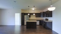 This single-floor space showcases a well-appointed kitchen as its centerpiece. The kitchen is designed with rich, dark-stained cabinetry that extends to the upper walls, providing ample storage throughout. Light-toned granite countertops offer a striking contrast against the dark cabinetry, creating a visually balanced aesthetic. A central kitchen island, finished with matching granite, serves as a functional workspace and gathering point, illuminated by two pendant lights that hang from the ceiling above.

Stainless steel appliances, including a refrigerator, dishwasher, microwave, and range, are seamlessly integrated into the cabinetry, contributing to a cohesive, modern look. The kitchen also features a sink positioned along the back counter wall. Dark hardwood flooring runs throughout, complementing the overall color scheme and adding warmth to the space. The light-colored walls provide a neutral backdrop that enhances the contrast between the dark cabinetry and flooring. The space feels open and functional, with a clean, contemporary design that balances practicality with an appealing visual character.