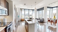 This thoughtfully designed two-bedroom, three-bathroom residence in McLean, Virginia offers a refined open-concept layout flooded with natural light. The living and dining areas are anchored by dramatic floor-to-ceiling windows that frame sweeping city skyline views, creating a bright and airy atmosphere throughout. Rich hardwood flooring flows seamlessly across the main living spaces, complementing the modern aesthetic.

The kitchen features dark cabinetry, stainless steel appliances, and a central island with pendant lighting, opening directly to the dining and living areas for effortless entertaining. The dining nook is wrapped in expansive corner windows, offering a sun-filled setting with outdoor views.

The primary bedroom provides a comfortable retreat, highlighted by a bold accent wall, plush carpeting, and direct access to a well-appointed en-suite bathroom. The bathroom includes a stone-topped vanity with dark wood cabinetry and a glass-enclosed walk-in shower with large-format tile. A dedicated walk-in closet with built-in shelving and hanging rods adds generous storage. The second bedroom mirrors the comfortable design of the primary. Three full bathrooms ensure convenience for residents and guests alike. This residence offers a polished urban living experience in the heart of McLean.