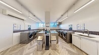 Located at 12-24 Leith Hill Rd in Toronto, this shared laundry facility is a thoughtfully designed, functional space that prioritizes both utility and comfort. The room is finished with a distinctive herringbone-patterned luxury vinyl flooring that adds a refined aesthetic touch throughout.

Multiple front-loading LG commercial washers and dryers are arranged along the walls, providing ample capacity for residents. A row of white shaker-style cabinetry with an integrated utility sink runs along one wall, offering generous counter space for folding and sorting laundry. A decorative dark tile backsplash accents the lower portion of the walls, complementing the clean white upper walls and bright, linear overhead lighting.

A waiting area is thoughtfully incorporated into the space, furnished with a black leather chair and a glass-top coffee table, allowing users to wait comfortably. A wall-mounted split air conditioning unit ensures year-round climate comfort. Natural light enters through windows positioned near the machines, enhancing the overall brightness of the room.

Informational notices and a corkboard bulletin board are mounted near the sink area, keeping users informed. The space is accessed through a glazed metal door, and a security camera is installed for added safety. Overall, this laundry facility is clean, modern, and well-appointed.