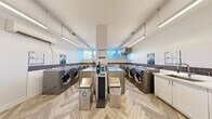 Located at 12-24 Leith Hill Rd in Toronto, this shared laundry facility is a thoughtfully designed, functional space that prioritizes both utility and comfort. The room is finished with a distinctive herringbone-patterned luxury vinyl flooring that adds a refined aesthetic touch throughout.

Multiple front-loading LG commercial washers and dryers are arranged along the walls, providing ample capacity for residents. A row of white shaker-style cabinetry with an integrated utility sink runs along one wall, offering generous counter space for folding and sorting laundry. A decorative dark tile backsplash accents the lower portion of the walls, complementing the clean white upper walls and bright, linear overhead lighting.

A waiting area is thoughtfully incorporated into the space, furnished with a black leather chair and a glass-top coffee table, allowing users to wait comfortably. A wall-mounted split air conditioning unit ensures year-round climate comfort. Natural light enters through windows positioned near the machines, enhancing the overall brightness of the room.

Informational notices and a corkboard bulletin board are mounted near the sink area, keeping users informed. The space is accessed through a glazed metal door, and a security camera is installed for added safety. Overall, this laundry facility is clean, modern, and well-appointed.