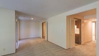 This single-floor space centers around a generously sized living area finished with wall-to-wall neutral carpeting and cream-toned walls that create a clean, understated backdrop. The textured popcorn ceiling adds a classic touch throughout. Louvered bifold doors on one side open to reveal the kitchen, which features dark cabinetry and hard-surface flooring, offering a functional contrast to the softness of the carpeted living space. A doorway on the adjacent wall leads to additional rooms, including what appears to be a space with a closet, suggesting a bedroom or utility area. Recessed lighting provides ambient illumination throughout. The open layout of the main living area allows for flexible furniture arrangement, while the clear sightlines between spaces contribute to an airy, connected feel. The overall design reflects a straightforward, functional aesthetic with a neutral palette suited to a variety of interior styles.