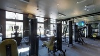 This contemporary fitness center offers a thoughtfully designed workout environment finished in a sleek, dark aesthetic. The space is outfitted with a full suite of professional-grade exercise equipment, including cable weight machines, treadmills, elliptical trainers, and a variety of strength training apparatus — providing everything needed for a complete fitness routine. Large floor-to-ceiling windows line the perimeter of the space, flooding the interior with natural light while offering open views of the surrounding outdoor environment. The ceiling features a combination of recessed lighting and suspended linear pendant fixtures, ensuring the space remains bright and well-lit throughout. Durable rubber flooring covers the workout area, providing both safety and comfort underfoot. A mounted flat-screen television is also present, adding an entertainment element to the experience. The overall ambiance is polished and modern, with the dark finishes on equipment and walls creating a focused, gym-quality atmosphere. The layout is efficiently organized, allowing for comfortable movement between stations and equipment throughout the training floor.