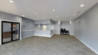 This single-floor clubhouse offers a clean, functional layout designed to serve as a central gathering space for residents and guests. The main area is open and spacious, featuring patterned carpet tiles in neutral grey tones that add texture while maintaining a professional aesthetic throughout. Soft blue-grey walls create a calm, welcoming atmosphere, complemented by bright recessed lighting in the white ceiling that ensures even illumination across the entire space. A prominent built-in reception or service counter anchors the central area, finished in white with a light-toned countertop. It separates the main open floor from a back area visible through a pass-through opening, suggesting utility or administrative use. A small cluster of black seating chairs is positioned along the far wall, providing a simple waiting or gathering area. The space is kept intentionally minimal and versatile, allowing for a variety of community uses such as meetings, events, or casual gatherings. The layout flows openly without visual clutter, offering flexibility in how the space can be arranged or utilized. Overall, the clubhouse presents a tidy, modern, and practical environment suited for community-focused activities.