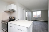 This single-floor unit offers a clean, modern aesthetic with a thoughtfully designed open-concept layout. The living area is spacious and bright, featuring dark wood-look laminate flooring, neutral grey walls, and a large window that allows natural light to fill the room. A wall-mounted sconce near the entryway adds a warm accent, while a baseboard heating unit and window-mounted air conditioning unit provide year-round climate control.

The kitchen is efficiently laid out with a galley-style design, featuring white cabinetry with sleek hardware, light-toned countertops, a white subway tile backsplash, and a full suite of stainless steel appliances including a refrigerator and electric range with overhead ventilation. Light wood-look flooring complements the kitchen's bright, clean finish.

The bedroom is a comfortable, well-proportioned room finished with plush grey carpeting and a large window offering outdoor views. Baseboard heating is also present, ensuring consistent comfort.

The bathroom is simply finished in white, featuring a tiled bathtub surround, clean fixtures, and brushed nickel towel bars that add a contemporary touch. Overall, the unit presents a modern, move-in-ready space with consistent finishes and a practical, functional floor plan.