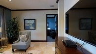 This single-floor space presents a refined interior characterized by a cohesive design language of dark espresso wood trim, warm neutral walls, and polished marble-look tile flooring. The main living area is furnished with a classic upholstered armchair accented with decorative throw pillows, positioned alongside a large flat-screen television mounted on the wall and supported by a rich wood media console. A framed abstract artwork adds a tasteful decorative element to the wall space. Crown molding and visible ceiling vents contribute to the room's structured, well-finished aesthetic. Through a dark-trimmed doorway, a dedicated office space is visible, distinguished by its deeper, moody blue-toned walls and darker ambiance. The office is equipped with a computer workstation, an ergonomic office chair, and track lighting overhead, making it a fully functional workspace. The transition between the two areas is defined clearly by the doorway framing, creating a sense of purposeful separation between relaxation and productivity zones. Overall, the space conveys a polished, professional atmosphere with consistent attention to architectural detail and interior finishing.