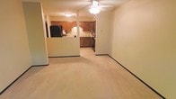 This single-floor residence offers a practical and comfortable layout centered around an open-concept living and kitchen area. The living space is finished with wall-to-wall neutral carpeting, creating a warm and inviting atmosphere, and is equipped with a ceiling fan that provides both lighting and air circulation. A low dividing half-wall separates the living area from the kitchen, maintaining an open feel while subtly defining each zone. The kitchen is well-appointed with rich, warm-toned wood cabinetry that runs along the walls, offering ample storage. Black appliances, including a full-size refrigerator and a gas or electric range, anchor the kitchen's functional design. The countertops provide a clean, usable workspace, and under-cabinet lighting adds a practical touch. Neutral-toned walls throughout the space lend a clean, understated aesthetic that is easy to personalize. The overall layout is efficient and cohesive, with the open sightlines between the living and kitchen areas making the space feel larger and well-connected. This single-level layout is ideal for those seeking a straightforward, easy-to-navigate floor plan with a comfortable, lived-in ambiance.