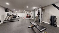 Located at Oxford House Apartments in Philadelphia, PA, this combined Fitness and Business Center is a thoughtfully designed amenity space that serves residents with both wellness and productivity needs. The fitness area features durable rubber flooring in a speckled gray finish, recessed lighting, and neutral gray walls that create a clean, modern atmosphere. Equipment includes True-brand treadmills, an elliptical trainer, a recumbent bike, a multi-station cable machine with attached weight rack, a adjustable weight bench, and a full set of dumbbells — providing a comprehensive range of cardio and strength training options. A large wall mirror spans one side of the gym, enhancing the sense of openness and allowing users to monitor their form. Seamlessly integrated into the fitness space, the business center is enclosed by sleek glass walls and doors, maintaining visual connectivity while offering a defined, quiet work environment. Inside, bold branding graphics of "Oxford House Apartments" adorn the walls, accompanied by two black desks, vibrant red ergonomic chairs, and computer workstations. A striking red, gray, and black patterned carpet anchors the business area with a pop of color and contemporary style. Together, these two functional zones form a cohesive, well-appointed amenity center designed for modern residential living.