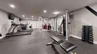 Located at Oxford House Apartments in Philadelphia, PA, this combined Fitness and Business Center is a thoughtfully designed amenity space that serves residents with both wellness and productivity needs. The fitness area features durable rubber flooring in a speckled gray finish, recessed lighting, and neutral gray walls that create a clean, modern atmosphere. Equipment includes True-brand treadmills, an elliptical trainer, a recumbent bike, a multi-station cable machine with attached weight rack, a adjustable weight bench, and a full set of dumbbells — providing a comprehensive range of cardio and strength training options. A large wall mirror spans one side of the gym, enhancing the sense of openness and allowing users to monitor their form. Seamlessly integrated into the fitness space, the business center is enclosed by sleek glass walls and doors, maintaining visual connectivity while offering a defined, quiet work environment. Inside, bold branding graphics of "Oxford House Apartments" adorn the walls, accompanied by two black desks, vibrant red ergonomic chairs, and computer workstations. A striking red, gray, and black patterned carpet anchors the business area with a pop of color and contemporary style. Together, these two functional zones form a cohesive, well-appointed amenity center designed for modern residential living.