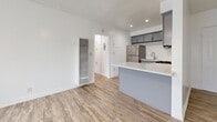 Located at 10237 Western Ave in Downey, California, this one-bedroom, one-bathroom unit offers a well-organized single-floor layout designed to maximize functionality. The kitchen is a standout feature, showcasing gray shaker-style cabinetry, a crisp white subway tile backsplash, stainless steel appliances including a refrigerator, gas range, and microwave, and a peninsula with a white quartz-style countertop that provides additional prep space and separates the kitchen from the living area. Wood-look laminate flooring runs consistently throughout the main living spaces, lending warmth and visual continuity. The open living area features clean white walls, recessed lighting, and a wall-mounted heating unit, creating a bright and functional atmosphere. A hallway connects the main living space to the bedroom and bathroom, with built-in cabinetry providing additional storage along the way. The bedroom is a comfortable, private retreat with light wood-look flooring, white walls, a textured ceiling, and a ceiling fan. The bathroom continues the clean aesthetic with light tile flooring and simple, modern finishes. Natural light filters in through windows visible in the 3D overview, enhancing the airy feel of the space throughout.