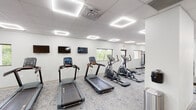 This single-floor fitness facility is thoughtfully designed to accommodate a wide range of workout preferences, offering distinct zones that flow seamlessly into one another across a spacious, open layout. The interior is finished with light-toned carpet throughout most areas, clean white and warm beige walls, and a suspended ceiling fitted with square LED panel lights that provide consistent, bright illumination across the entire space.

The cardio zone is well-appointed with multiple treadmills, elliptical trainers, a stationary bike, and an air bike, complemented by wall-mounted flat-screen televisions. Windows along the perimeter allow natural light to filter in and offer views of the surrounding greenery. The strength training area features a cable machine, weight benches, dumbbell racks, and a multi-functional training rig equipped with suspension straps, medicine balls, kettlebells, and a heavy bag — ideal for functional and high-intensity training.

A dedicated functional training zone includes a branded performance mat and agility runner, providing space for dynamic movement and conditioning. An adjacent glass-partitioned room, visible from the main floor, appears suited for group sessions or private use. Direct exterior access via glass and solid doors adds convenience to this clean, modern facility.