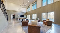 Located at 2920 Frontage Rd in Gainesville, Georgia, this impressive amenity space is defined by its dramatic double-height ceilings that create an expansive, open atmosphere throughout the main floor. Vertical shiplap-style wall paneling in a warm sage tone runs the full height of the interior, lending a refined yet casual character to the space.

An abundance of natural light pours in through a generous array of large, floor-to-ceiling and clerestory windows lining the exterior wall, offering clear views of an outdoor patio and surrounding wooded landscape. The polished concrete floors reflect this natural light, further enhancing the bright and airy feel.

A striking circular chandelier serves as a central focal point overhead, adding an elegant touch to the industrial-inspired design. The lounge area features plush, tufted leather sofas and armchairs arranged on a layered area rug, creating a comfortable gathering space. A wall-mounted flat-screen television anchors one end of the room, complemented by decorative wall art and a coordinating storage console.

A full-sized JOOLA ping-pong table occupies a prominent position on the open floor, offering recreation for visitors. A beautifully crafted iron-railed staircase leads to an upper mezzanine level, adding architectural interest and vertical dimension. The overall ambiance is modern, welcoming, and well-suited for social and recreational use.